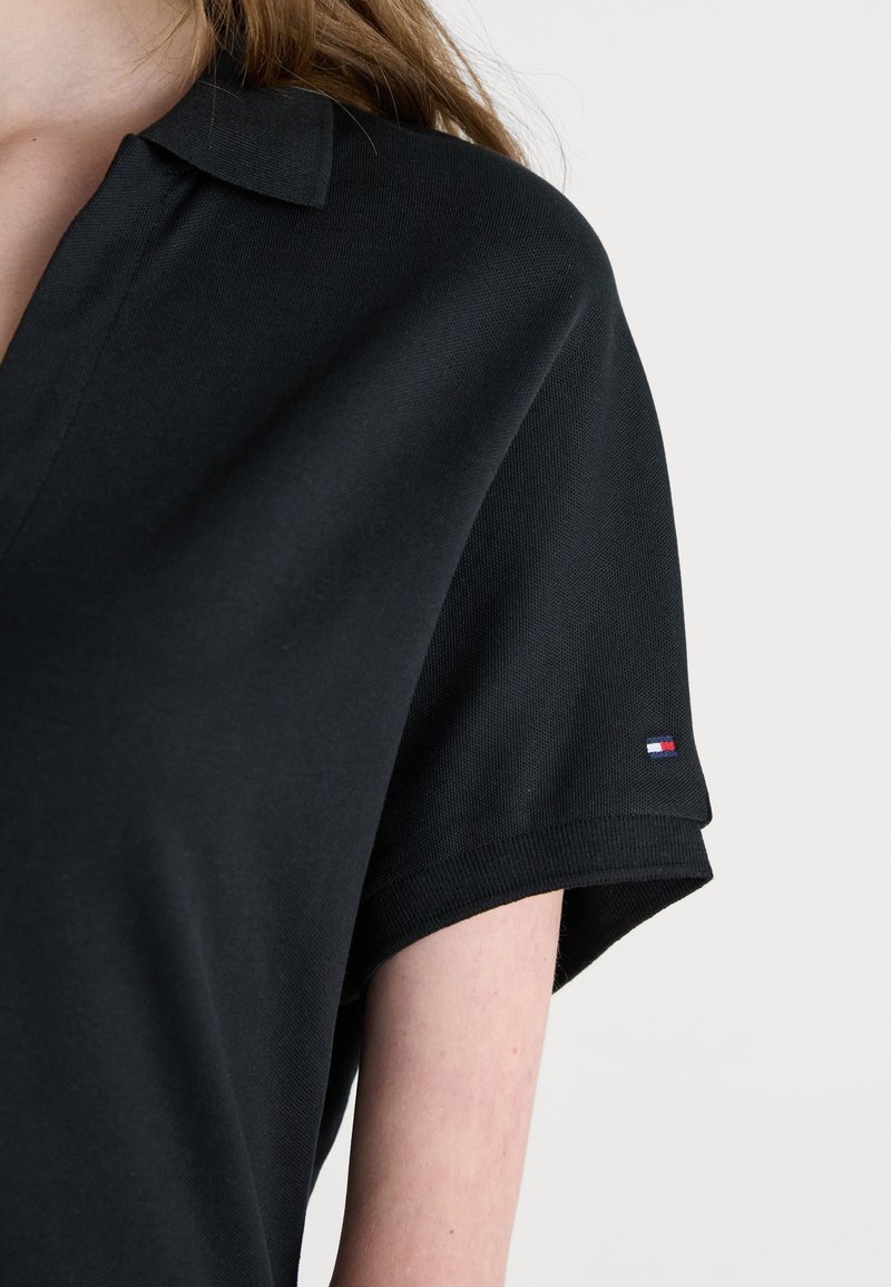 Polo noir en tissu texturé, avec un col classique et des manches courtes. Logo brodé notable sur le poignet.