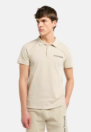 Ung mand iført beige poloshirt og matchende bukser med "bruno banani" logo, står mod en ensfarvet baggrund og kigger mod venstre.