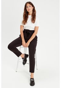 Schwarze hochtaillierte Hosen mit einer Schleifendetail an der Taille, kombiniert mit einem kurzärmeligen weißen Oberteil und schwarzen Mokassins mit einer Kettenakzent.