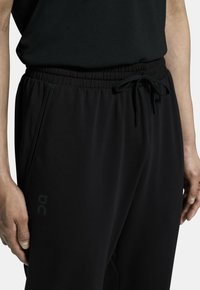 Pantalons de sport noirs avec une taille élastique et un cordon de serrage ajustable. Comprennent des poches latérales et un logo discret sur la hanche. Texture de tissu douce.