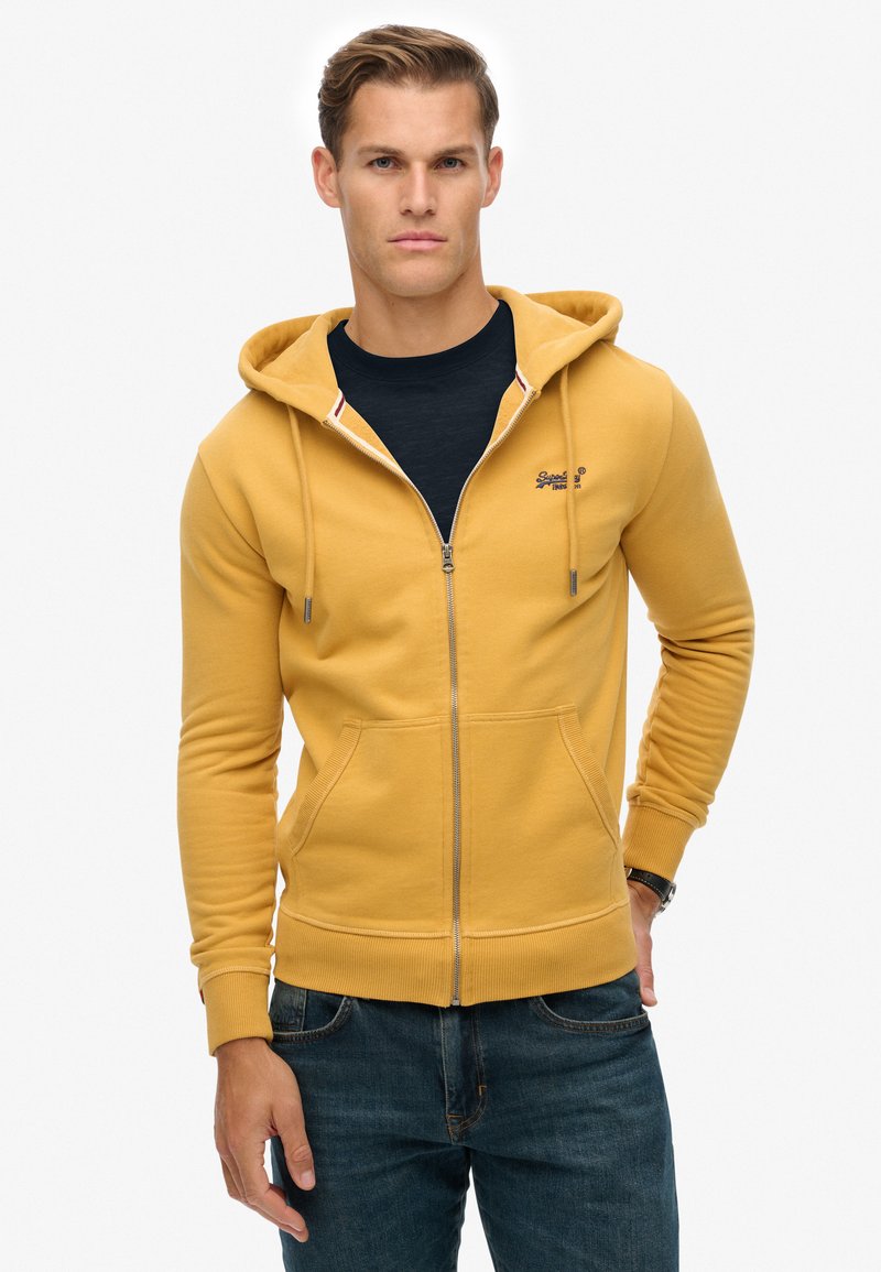 Uomo che indossa un hoodie giallo senape con zip sopra una maglietta nera, abbinato a jeans blu, in piedi contro uno sfondo bianco semplice.