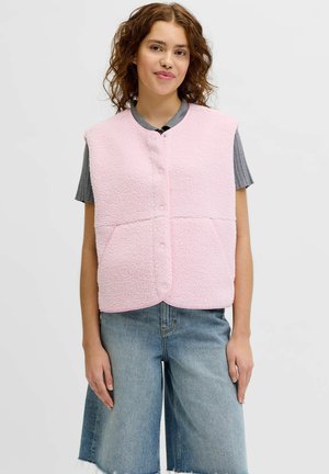 Ung kvinde iført en lyserød fleecevest med knapper over en grå kortærmet top og løstsiddende blå cowboyshorts.