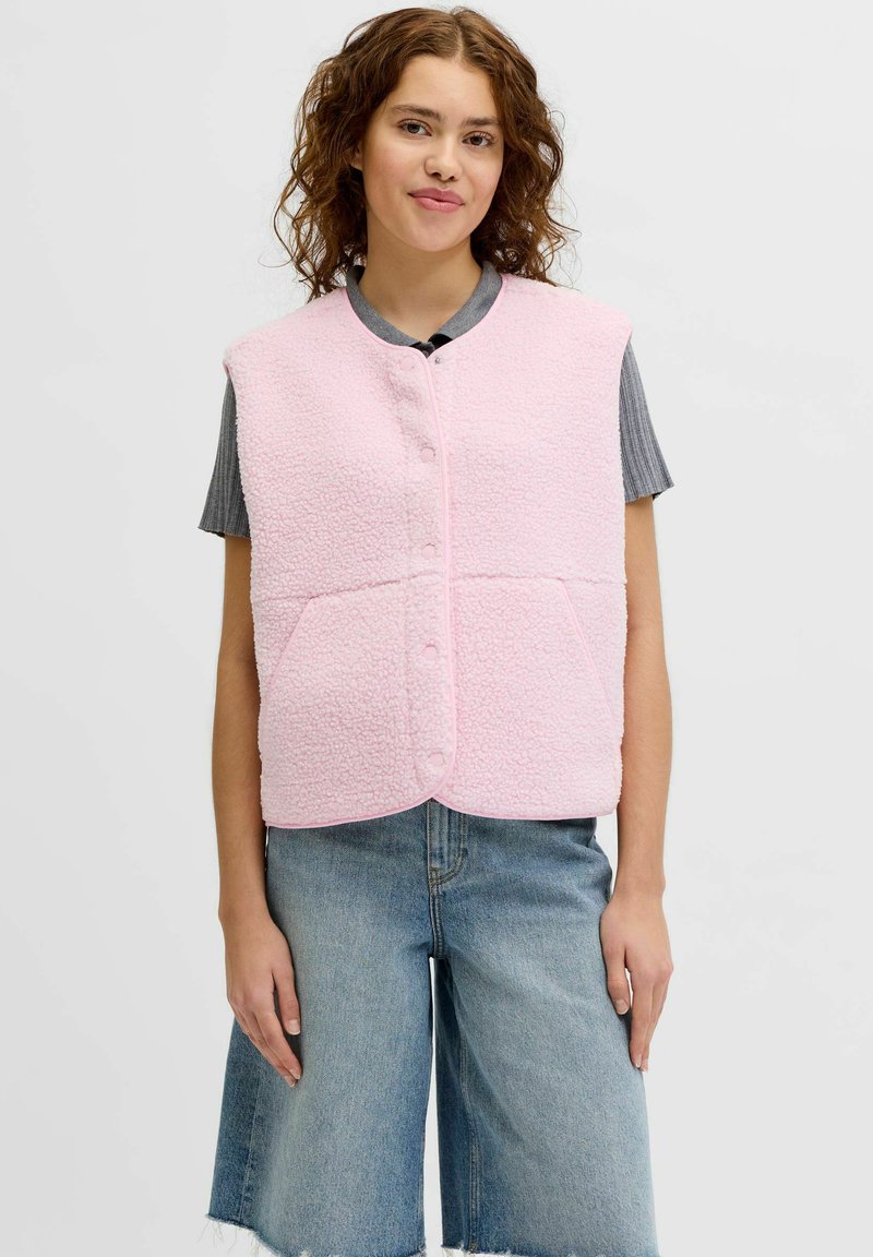 Ung kvinde iført en lyserød fleecevest med knapper over en grå kortærmet top og løstsiddende blå cowboyshorts.