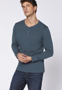 Langärmliges, teal-farbenes Henley-Shirt mit drei Knöpfen, rundem Halsausschnitt und dezentem gesticktem Logo auf der Brust, kombiniert mit dunklen Jeans.