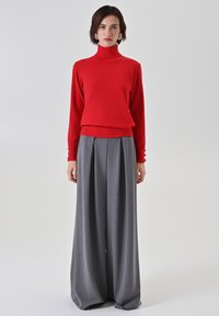 Roter Rollkragenpullover mit langen Ärmeln, kombiniert mit einer hochgeschnittenen, weit geschnittenen grauen Hose mit Falten und Knopfakzenten an den Ärmeln.