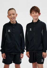 Två leende barn i svarta Hummel sportjackor och shorts, stående sida vid sida mot en vit bakgrund.