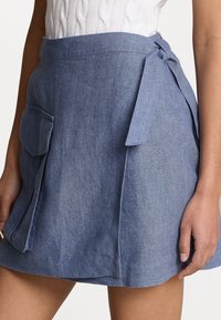 Gonna in denim blu chiaro, con un fiocco in vita, due tasche frontali e una vestibilità rilassata con un orlo leggermente svasato.