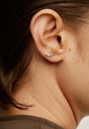 Primer plano de una oreja derecha con cabello corto castaño que lleva un pequeño pendiente de oro con tres piedras redondas dispuestas en forma triangular.