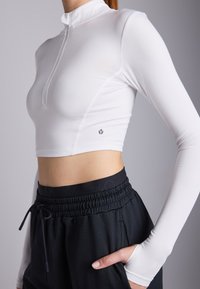 Bijela dugački rukav kratak top s polu-zip ovratnikom. Ima glatku tkaninu i logotip u donjem lijevom kutu. Uparen s crnim hlačama elastičnog struka.
