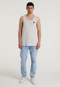 Šviesiai pilkas tank topas su apvalia iškirpte ir mažuoju juodu logotipu. Dėvima su šviesiai mėlynomis džinsinėmis kelnėmis ir baltais sportbačiais. Lygus audinys, laisvas siluetas.