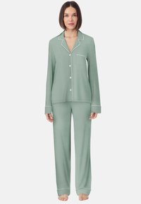 Vrouw die blootsvoets staat en een lichtgroen lange mouwen pyjama-attribuut met witte piping en borstzak draagt, tegen een effen achtergrond.