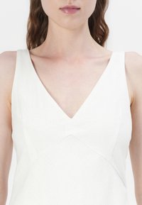 Weißes Leinenkleid mit V-Ausschnitt, tailliertem Oberteil und diagonalen Nahtdetails. Leichter und strukturierter Stoff, der ein minimalistisches Design zeigt.