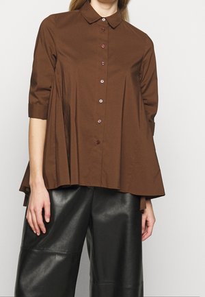 Kvinde iført en løs brun skjorte med trekvartlange ærmer og brede sorte læderbukser mod en ensfarvet baggrund.