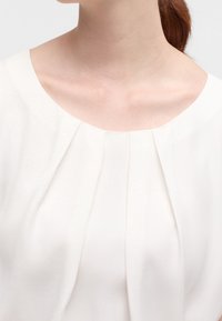 Vit blus med rund halsringning och vertikala veck framtill, med ett slätt, lätt tyg och en mjuk textur.