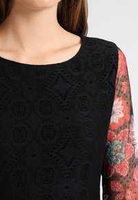 Vestido negro con cuerpo de encaje texturizado y mangas transparentes con estampado floral en rojo, rosa y verde azulado. Escote redondeado y diseño de tela detallado.