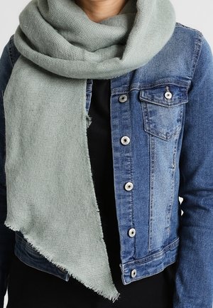 Personne portant une écharpe tricotée gris clair enroulée autour du cou et une veste en jean bleue avec des boutons argentés par-dessus un haut noir.