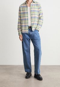 Koszula zapinana na guziki w kratę w pastelowych odcieniach zieleni, żółci i niebieskiego; zestawiona z luźnymi niebieskimi dżinsami i czarnymi butami z łańcuszkowym zdobieniem.