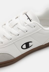 White leather sneaker with round toe, flat lace-up design, black embroidered logo, and a brown rubber sole. Textured surface details present.