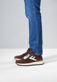 Brązowe zamszowe trampki z białymi akcentami, okrągłym noskiem i grubą beżową podeszwą. Noszone z niebieskimi dżinsami, prezentujące swobodny styl.