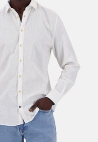 Weißes Leinen-Hemd mit langen Ärmeln, ausgestattet mit hellbraunen Knöpfen und einem kontrastierenden Kragen, getragen zu blauen Denim-Jeans.