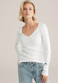 Valge pika varrukaga ribakudumiga henley särk V-kaelusega, kolm nuppu rinnal ja tihkenenud varrukad, sobitatud helesiniste teksadega.