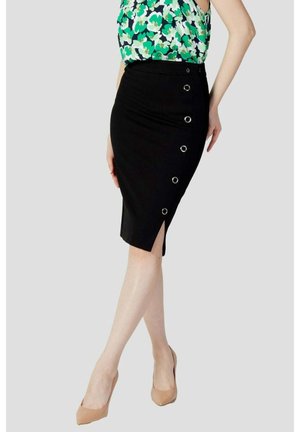 Kobieta ubrana w czarną ołówkową spódnicę z bocznymi srebrnymi oczkami oraz zielony kwiecisty top bez rękawów, w połączeniu z beżowymi szpilkami.