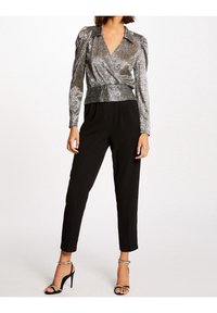 Eine Frau trägt eine silberne metallische Wickelbluse, schwarze taillierte Hose und schwarze High-Heels-Sandalen mit Knöchelriemen, während sie vor einem weißen Hintergrund steht.