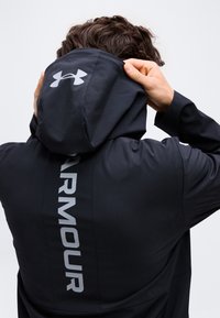 Άτομο με μαύρο μπουφάν Under Armour με την κουκούλα ανασηκωμένη, ρυθμίζει την κουκούλα με τα δύο χέρια, εμφανές λογότυπο και κείμενο "ARMOUR" στην πλάτη.