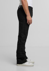 Muž stojaci bokom na jednoduchom šedom pozadí v čiernych rovno strihaných nohaviciach a bielych teniskách.