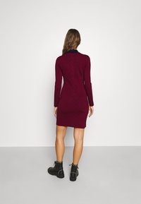 Robe longue manches longues couleur bordeaux avec une silhouette ajustée et un motif subtil. Dotée d'un col montant, elle est associée à des bottines noires à lacets.