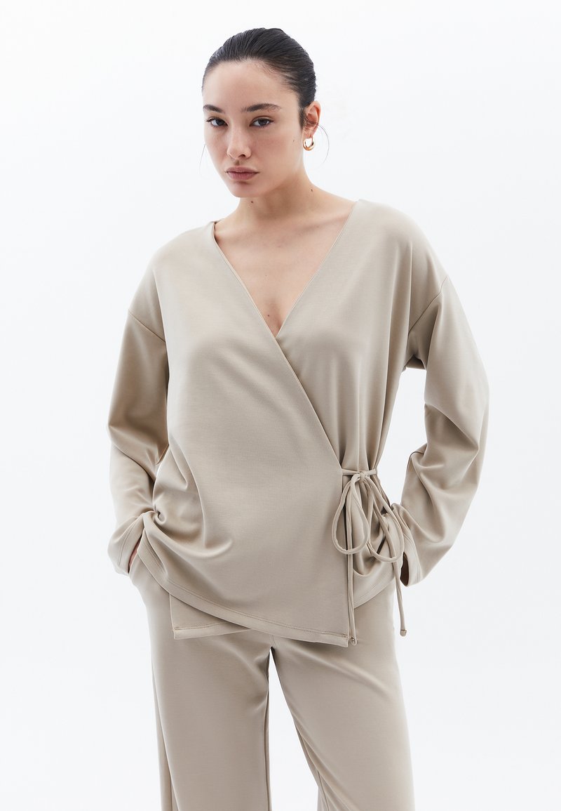 Beige wikkeltop met lange mouwen, v-hals en zijstrikdetail. Gemaakt van een gladde stof, gecombineerd met bijpassende beige broek.