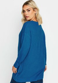 Överdimensionerad blå stickad tröja med ribbad textur och långa ärmar, med sidofickor och en lös passform, bärs över mörka jeans.