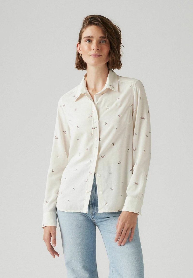 Camicia bianca a maniche lunghe con piccoli motivi floreali e colletto classico. Abbinata a jeans di denim azzurro chiaro.