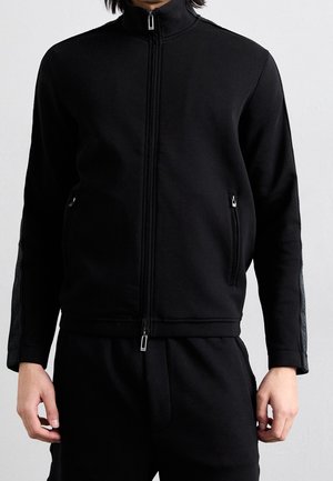 Person trägt eine schwarze Jacke mit Reißverschluss und hohem Kragen, vordere Reißverschlusstaschen und passende schwarze Hose, steht vor einem einfarbigen hellen Hintergrund.