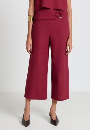 Donna che indossa pantaloni bordeaux a vita alta e gamba larga con top abbinato, cintura larga in tessuto, bracciale rigido dorato e tacchi neri a punta.
