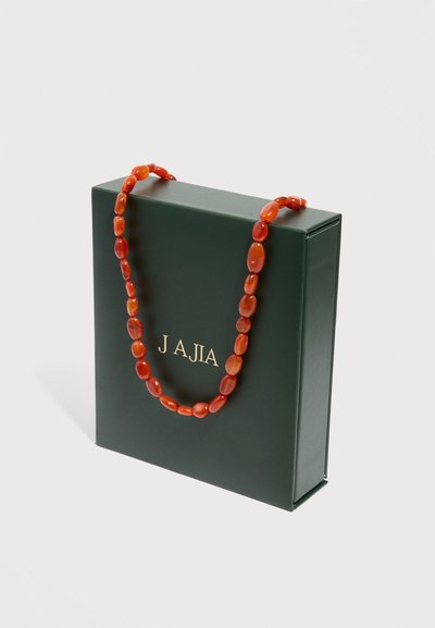Boîte cadeau vert foncé avec le texte doré "JAJA" et un collier de perles rouges drapé sur le dessus, sur un fond blanc uni.