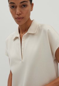 Beige Polohemd mit kurzen Ärmeln, V-Ausschnitt und weicher Textur. Verfügt über einen klassischen Kragen und subtile Nähte.