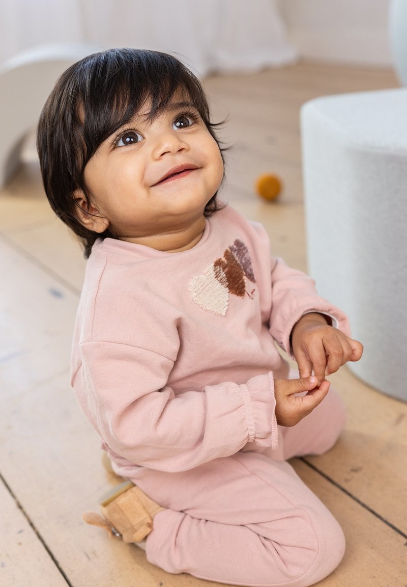 Lichtroze sweatshirt en broek, met een hartontwerp. Zachte, gestructureerde stof met gerimpelde boorden. Kind zit op een houten vloer.