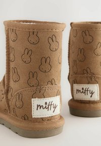 Bruna mockaskor med tryck av Miffy-kaniner över hela skon, texturerad yta, mjukt inre samt en tygetikett med "miffy" broderad.