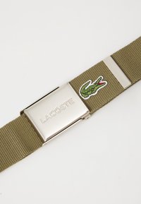 Homoszínű szövött öv ezüst fém csattal, amelyen a "LACOSTE" felirat van gravírozva, valamint zöld hímzett krokodil logó piros részletekkel.