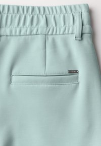 Pantalon bleu clair avec une taille élastique, texture lisse et une poche arrière unique avec une étiquette argentée.