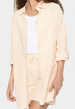 Woman wearing peach and white vertical-striped button-up shirt with rolled sleeves, matching shorts with drawstring, and white tank top.