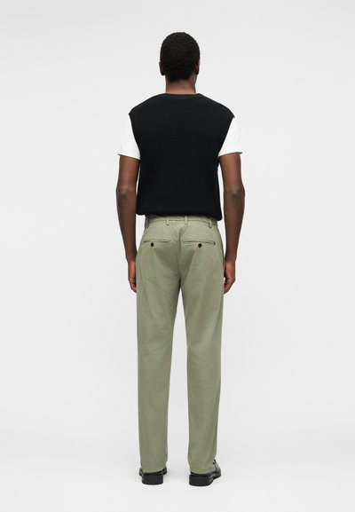 Άνδρας φορώντας μαύρο πλεκτό γιλέκο χωρίς μανίκια πάνω από λευκό κοντομάνικο πουκάμισο, ανοιχτό πράσινο παντελόνι και μαύρα παπούτσια, στέκεται γυρισμένος με την πλάτη.