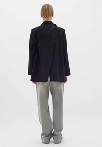 Črn blazer z edinstveno zasnovo na hrbtni strani, lahka tkanina, dolgi rokavi, kombiniran s svetlo sivo ravnimi hlačami. Gladka tekstura, minimalističen stil.