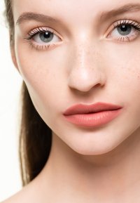 Närbild av ett ansikte som visar slät hud, lätta fräknar, fylliga läppar med matt persikofärgad läppstift och klara blå ögon. Naturligt sminkutseende.