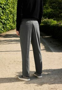 Szare spodnie o luźnym kroju, z tylnymi kieszeniami, zestawione z szarymi butami typu slip-on na żwirowej ścieżce wśród zieleni.