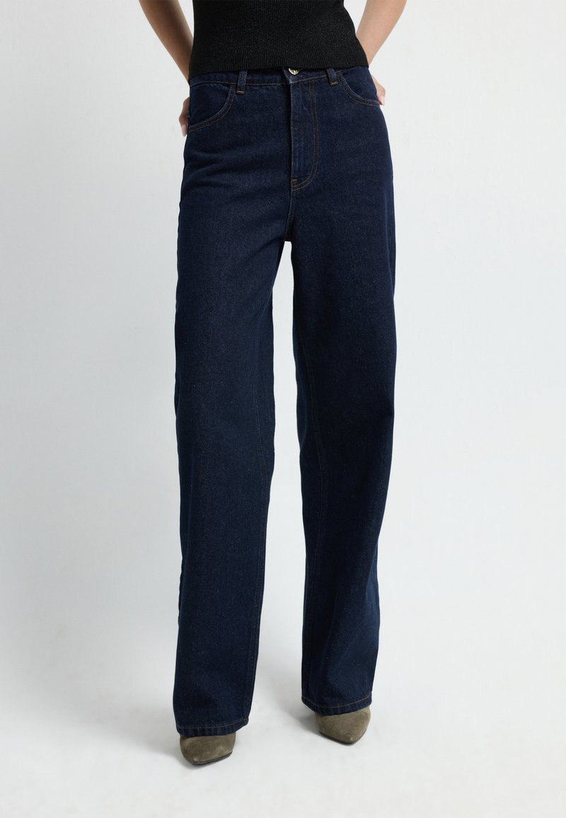 Granatowe dżinsy szerokie z nogawkami wykonane z denimu, z wysokim stanem, przednimi kieszeniami i kontrastowym przeszyciem. Modelka ma na sobie botki za kostkę.