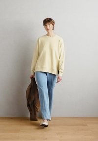 Felpa gialla a girocollo sopra jeans di denim blu chiaro, abbinata a una giacca marrone appoggiata su un braccio e ballerine nere su un pavimento di legno.