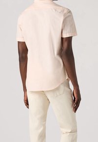 Camisa de algodón color rosa claro de manga corta con cuello, dobladillo curvado y detalle de pinza en la espalda, combinada con pantalones de color crema.