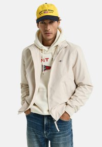 Beige zip-up jas, hoodie met logo, blauwe jeans en een gele en marineblauwe pet met tekst. Soepele textuur, casual ontwerp.